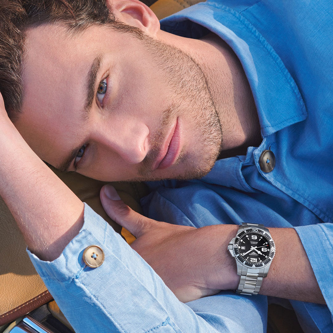 A man in a light blue button-up shirt reclines on a brown surface, gazing at the camera. On his left wrist, he wears a Longines Hydroconquest Automatic 41mm Watch with a black dial and unidirectional rotating bezel.