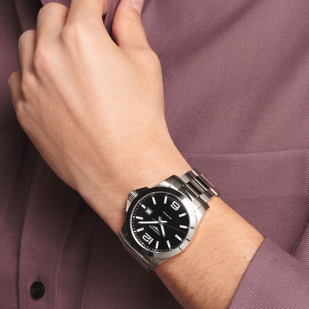 A person wearing a mauve long-sleeve shirt sports a Longines Conquest Quartz 43mm Watch by Longines, its stainless steel case and black dial with silver markers adding sporty elegance as their hand rests near the chest.