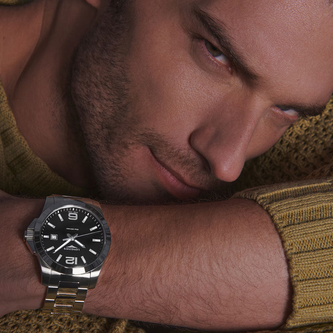 A man with light facial hair and blue eyes, in a gold knit sweater, leans his head on his arm while wearing the Longines Conquest Quartz 43mm Watch—a sporty yet elegant timepiece with a silver case and black dial.