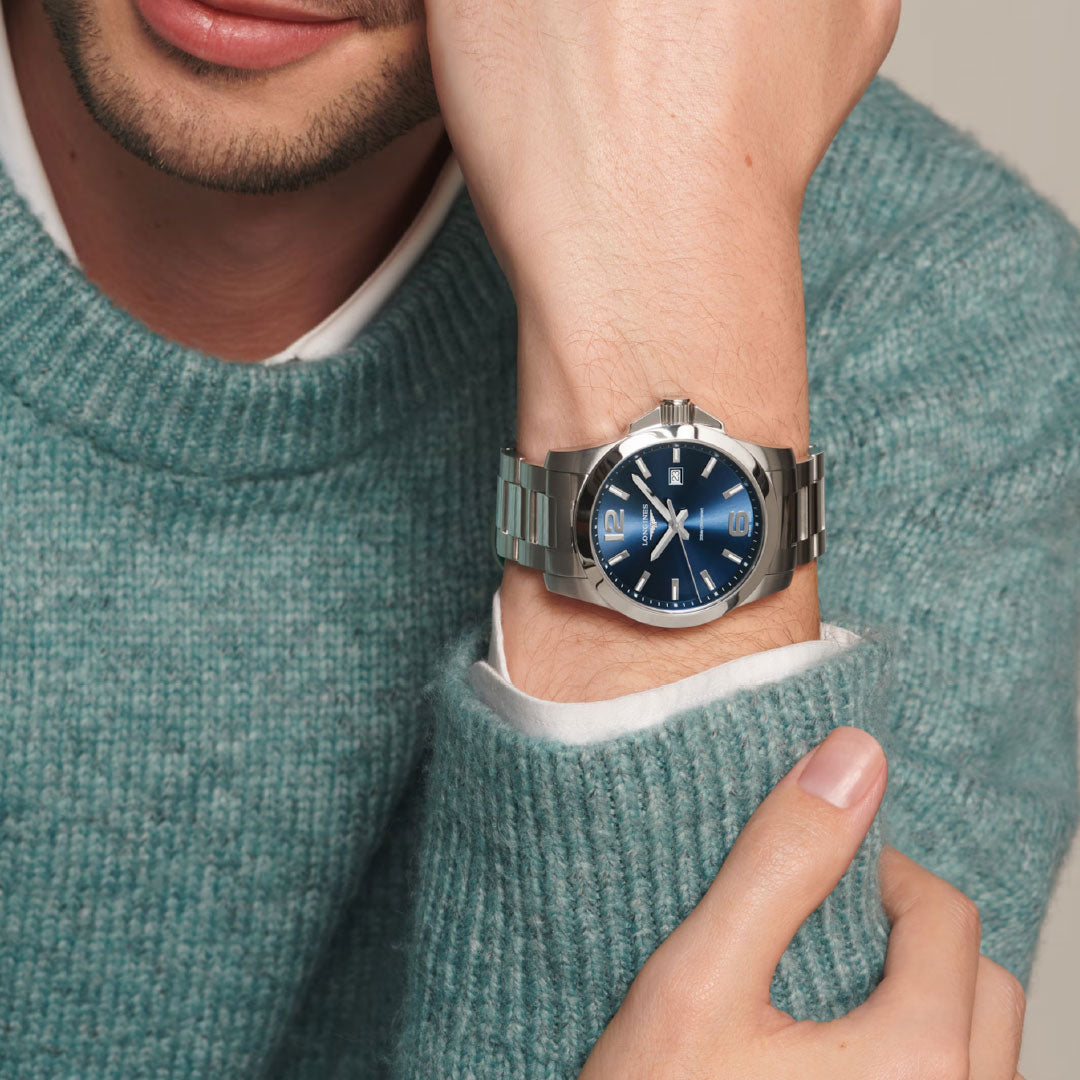 A person in a teal knit sweater rests their chin on their hand, showcasing the Longines Conquest Quartz 43mm Watch, which features a blue dial and silver hour markers. The luxury watch and the person's relaxed pose are highlighted.