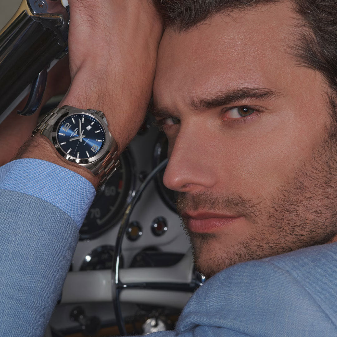 A man in a light blue suit poses inside a car, resting his head on his arm and showcasing the Longines Conquest Quartz 43mm Watch by Longines with a blue face. The car’s dashboard and steering wheel appear in the background.
