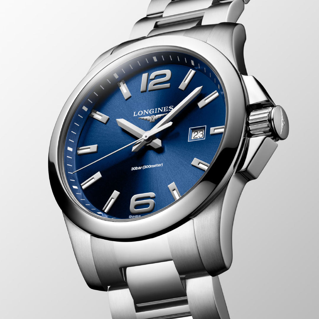 A close-up of the Longines Conquest Quartz 43mm Watch shows its blue dial, silver hour markers and hands, date window at 3 o’clock, and stainless steel bracelet against a light gradient background.