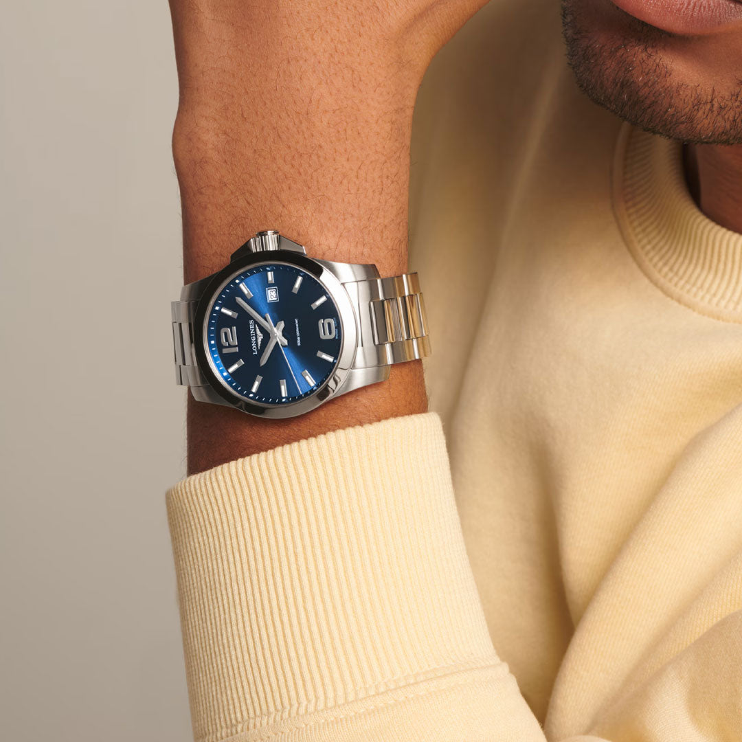 A person in a light yellow sweatshirt rests their chin on their hand, featuring a Longines Conquest Quartz 43mm Watch—a stylish silver wristwatch with a blue face and metal band—on their wrist.