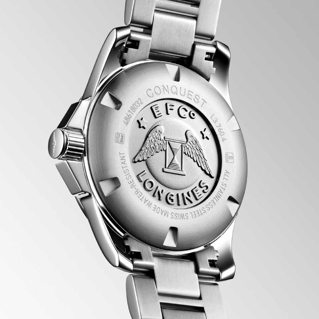 The image shows the back of a Longines Conquest Quartz 43mm Watch with a brushed stainless steel case and bracelet, engraved winged hourglass logo, and decorative text.