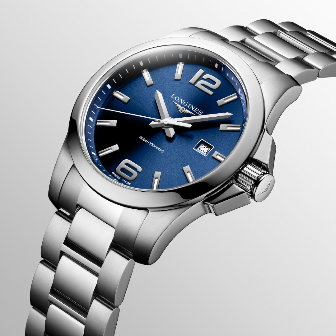 The Longines Conquest Quartz 43mm Watch features a blue dial, silver hour markers, and a date window at 3 o'clock. Styled in silver and set against a light gray background, it’s an elegant everyday luxury from Longines.