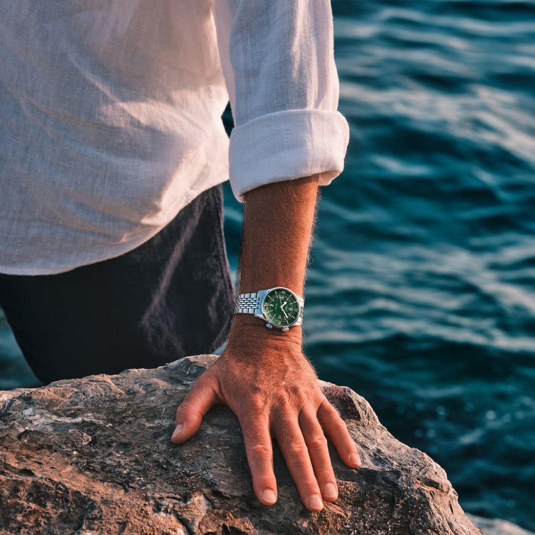 A person wearing a long-sleeve white shirt and a Longines Legend Diver Automatic 39mm Watch with a green dial rests their hand on a rock by the ocean, against the calming backdrop of deep blue water.