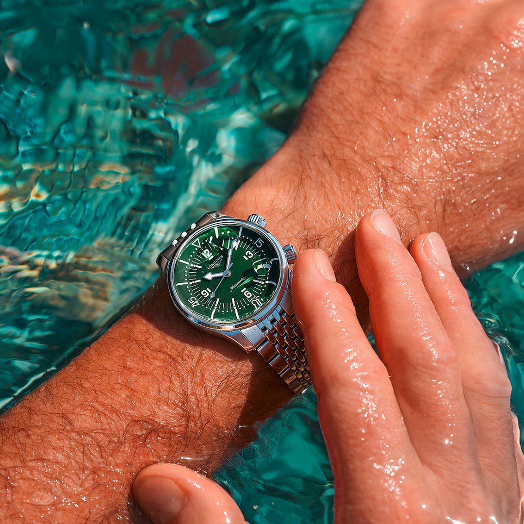 A wrist adorned with a Longines Legend Diver Automatic 39mm watch, featuring a green face, is submerged in clear, turquoise water. A hand with slightly wet skin rests gently over the wrist, highlighting this Longines watch's reflective surface in sunlight.