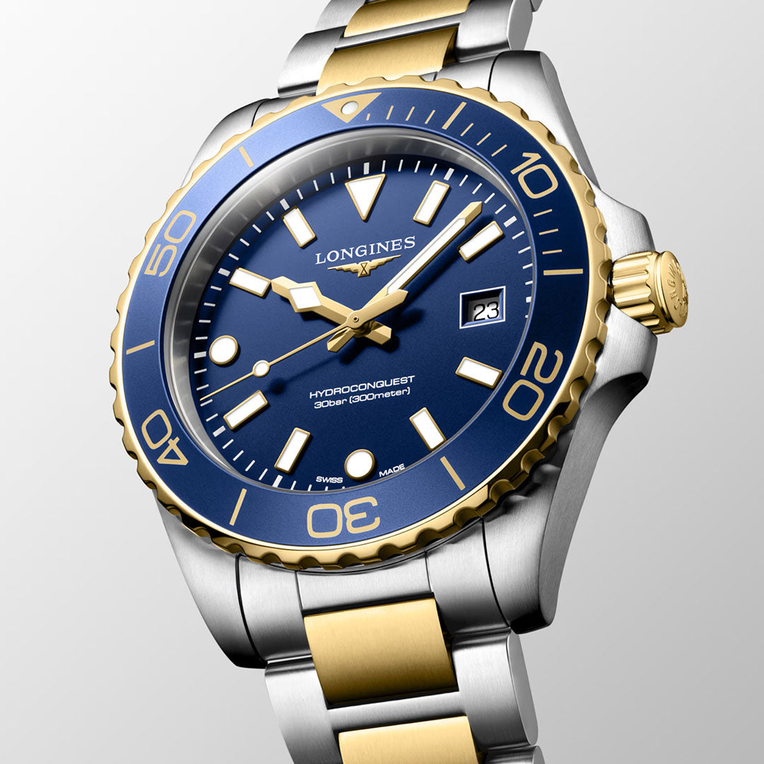 A close-up of the Longines HydroConquest Quartz 42mm Watch by Longines, featuring a blue dial with gold accents, a date display, and a two-tone silver and gold metal bracelet on a light gray background—an elegant representation of Swiss precision in luxury sport watches.