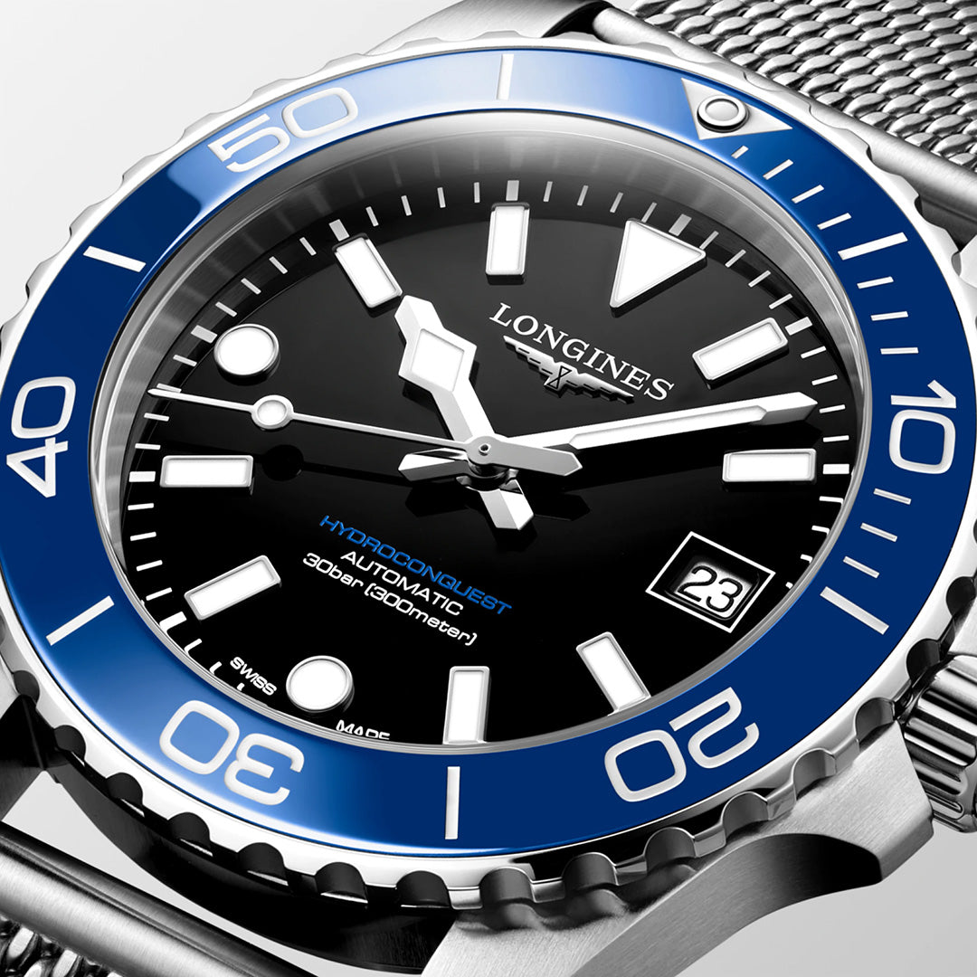 A detailed shot of the Longines HydroConquest Automatic 39mm Watch from Longines’ acclaimed sport collection, showcasing a blue bezel, black dial, silver hour markers, and a date window at 3 o’clock. This Swiss timepiece is paired with a stainless steel mesh strap.