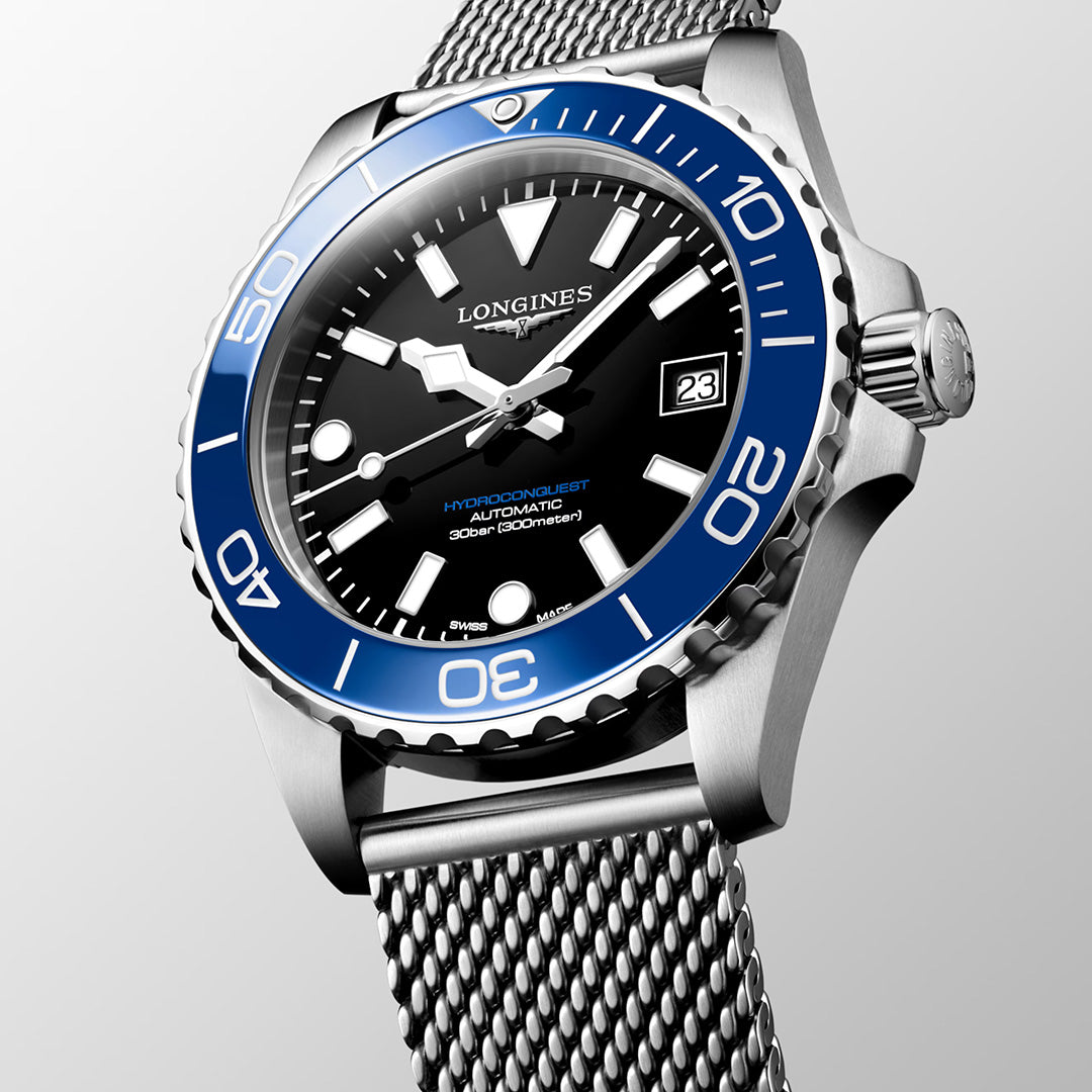 A Longines HydroConquest Automatic 39mm Watch by Longines, featuring a blue bezel, black dial, date window at 3 o’clock, and a stainless steel mesh bracelet from the sport collection, is displayed against a light gray background.
