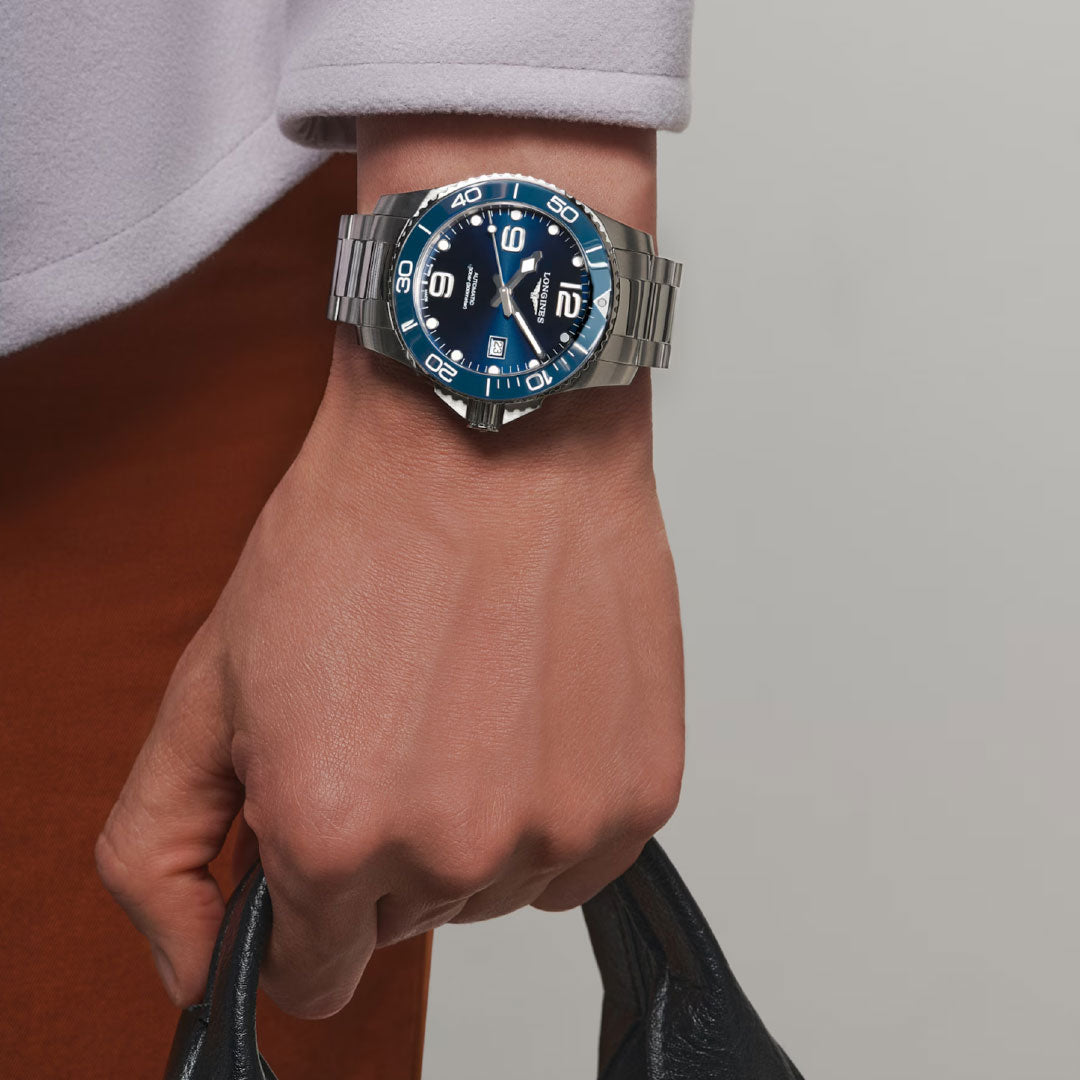 A person wearing a Longines Hydroconquest Automatic 43mm watch in blue and silver holds a black textured bag, dressed in a white sleeve and brown pants; the Longines watch face shows 10:10.