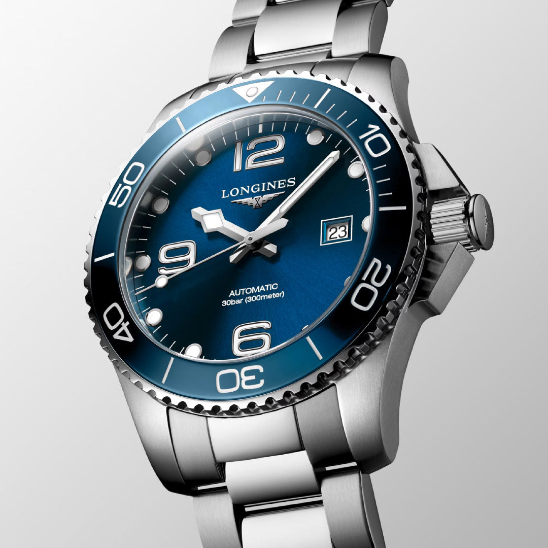 A close-up of the Longines Hydroconquest Automatic 43mm Watch, featuring a blue dial, silver hour markers, date display at 3 o’clock, stainless steel bracelet, and water-resistant design by Longines against a light gray background.
