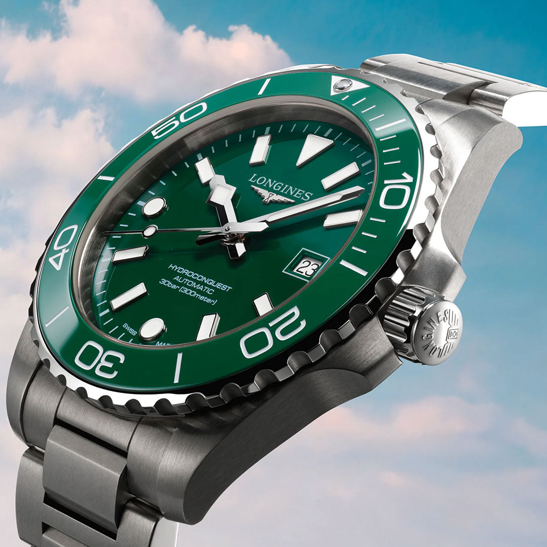 A close-up of the Longines Hydroconquest Automatic 42mm Watch featuring a green bezel and dial, silver hour markers, and a stainless steel bracelet, set against a blue sky with pink clouds.