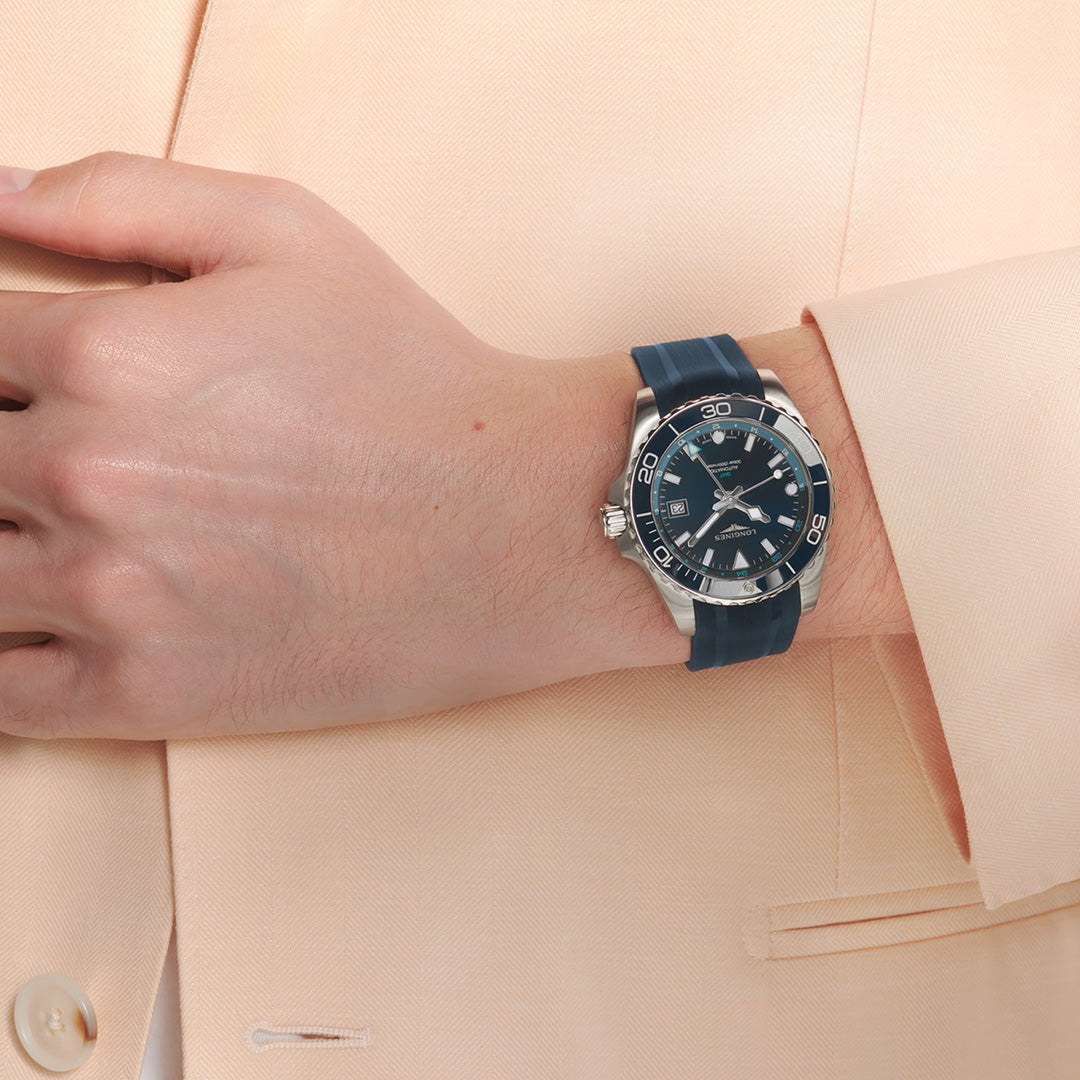 A person dressed in a light beige suit reveals their wrist adorned with the Longines HydroConquest GMT Automatic 41mm Watch, showcasing a striking blue and silver design from Longines. The watch features a dark dial illuminated by Swiss Super-LumiNova and is finished with a comfortable rubber strap.