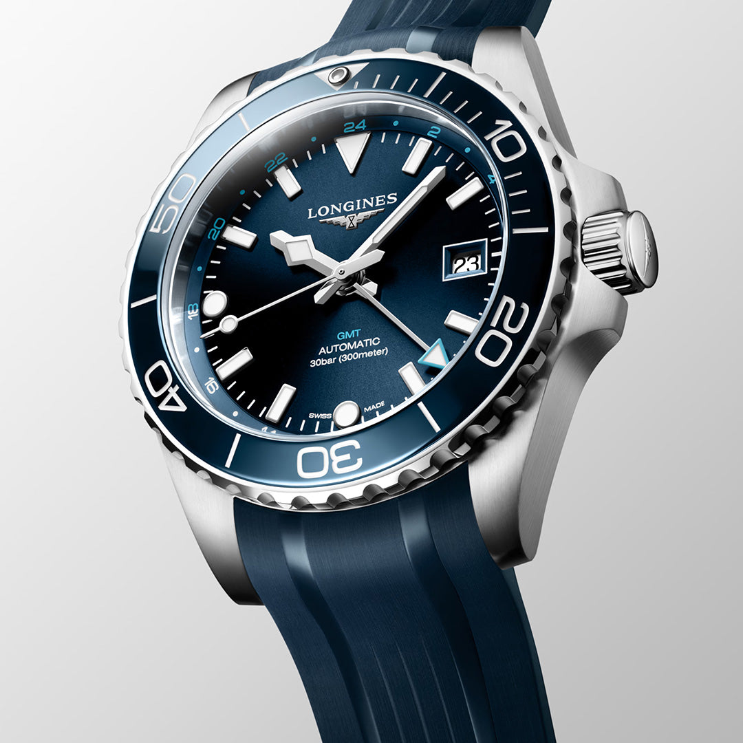 The Longines HydroConquest GMT Automatic 41mm Watch, from the esteemed Longines Sport Collection, is presented at an angle on a light background. This watch showcases a blue dial with Swiss Super-LumiNova accents, a date display at 3 o’clock, a silver bezel, and is paired with a blue rubber strap.