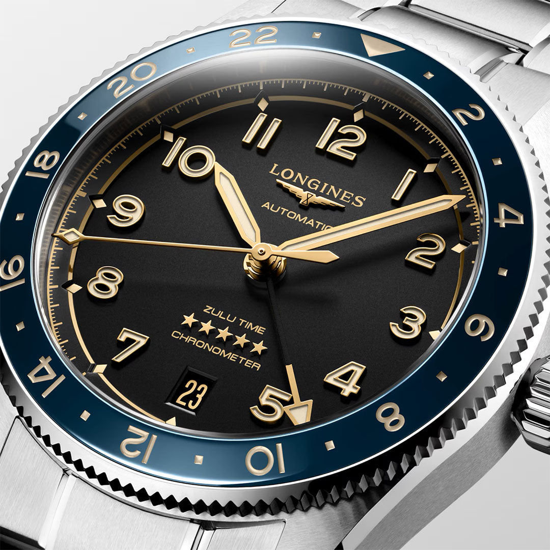 Close-up of the Longines Spirit Zulu Time Automatic 39mm Watch by Longines, featuring a black dial, gold hands and numerals, chronometer-certified automatic movement, a date window at 6 o’clock, and a blue and silver bezel with 24-hour markings for dual time zone functionality.