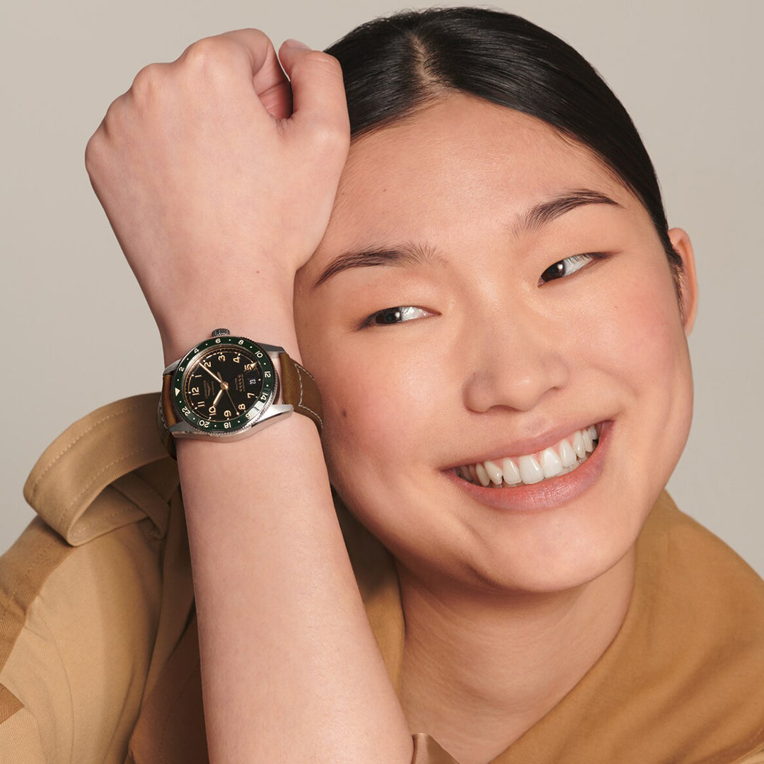 A person with straight black hair smiles, resting their head on their arm, wearing a Longines Spirit Zulu Time Automatic 39mm Watch. The sleek green and silver design with a ceramic insert complements their light brown shirt, effortlessly embodying dual time zone elegance.