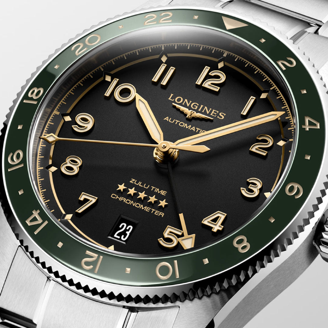 A close-up of the Longines Spirit Zulu Time Automatic 39mm Watch by Longines, featuring a black dial with gold numerals and hands, green 24-hour bezel, date at 6 o’clock, and stainless steel bracelet.