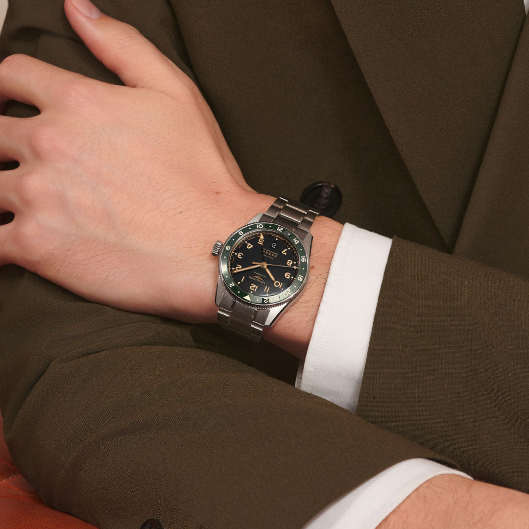 A person in a brown suit with visible white shirt cuffs crosses their arms, wearing the Longines Spirit Zulu Time Automatic 39mm watch by Longines, featuring a black and green bezel and a dark dial.