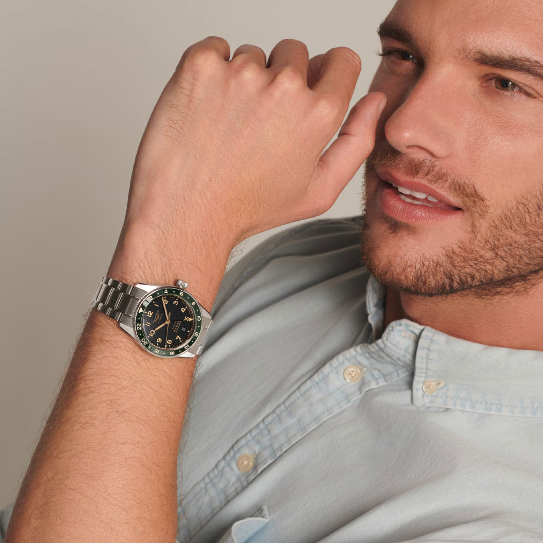 A man with trimmed facial hair, in a light blue button-up shirt, poses with his hand near his mouth, showcasing the Longines Spirit Zulu Time Automatic 39mm Watch by Longines, featuring a black and green bezel on his wrist.