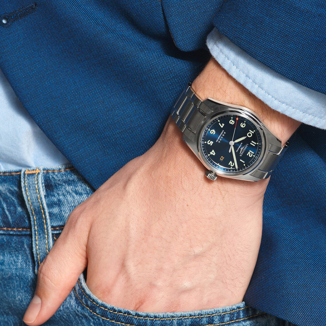 Close-up of a hand in blue jeans' pocket with a Longines Spirit Automatic 40mm Watch featuring a black dial and white numerals. A partially visible blue blazer sleeve suggests a casual yet stylish vibe.