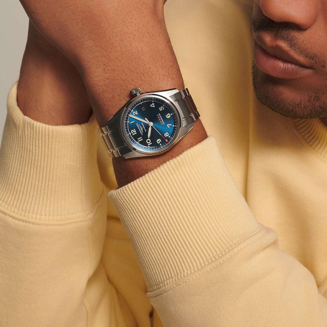 A person in a light yellow sweater rests their chin on their hand, showcasing a Longines Spirit Automatic 40mm Watch. The silver watch features a blue dial, silver hour markers, and a metal link band. The person is visible from the nose down.
