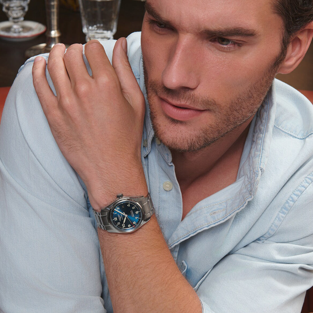 A man in a light blue shirt rests his arms on a table, featuring the Longines Spirit Automatic 40mm Watch with a silver bezel and blue dial on his left wrist. Blurred glassware is visible in the background.