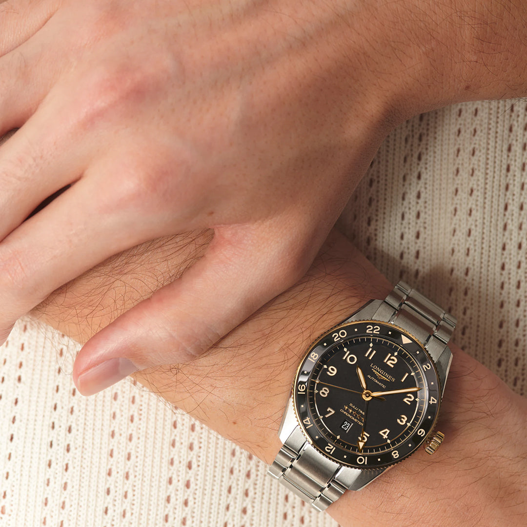 A person with a Longines Spirit Zulu Time Automatic 42mm watch in silver, featuring a black dial and silver hour markers, rests their hand on beige knit fabric, emphasizing the dual time zone function of this chronometer-certified piece.