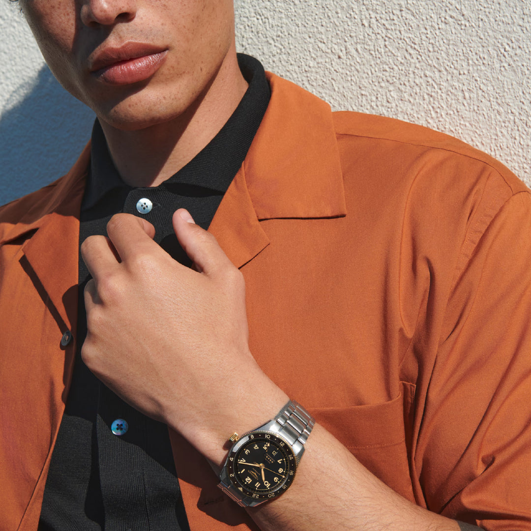 An individual in an orange shirt and black polo displays a silver Longines Spirit Zulu Time Automatic 42mm watch against a textured white wall. This chronometer-certified watch, featuring dual time zones, boasts a black dial with gold accents for a stylish yet casual look.