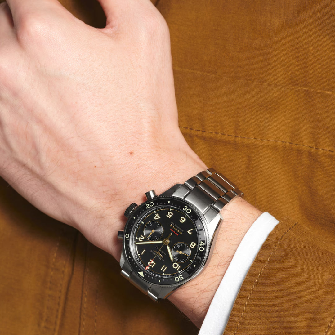 A close-up of a wrist displays the Longines Spirit Flyback Titanium Automatic 42mm Watch, featuring a striking silver and black design with a metal band. The black dial, adorned with subdials, highlights precision and elegance over a brown jacket sleeve.