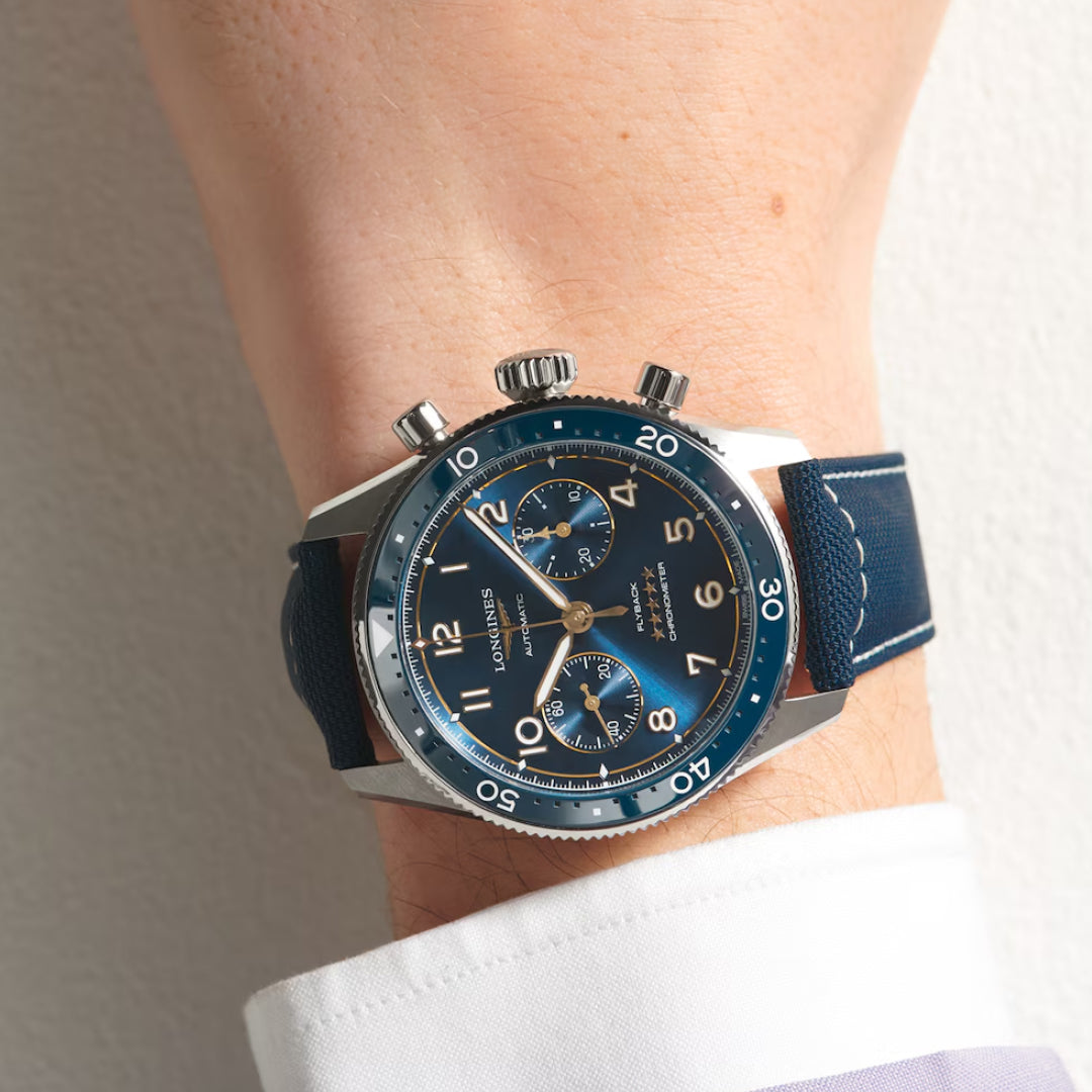 A close-up shows a wrist wearing the Longines Spirit Flyback Automatic 42mm Watch. It features a blue dial and strap, large face, three sub-dials, and silver hour markers. This chronometer-certified timepiece pairs perfectly with the wearer's crisp white shirt.