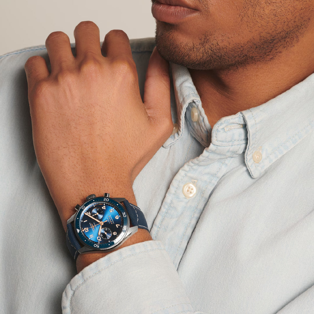 A person wearing a light blue button-up shirt rests their hand on their chin, showcasing the Longines Spirit Flyback Automatic 42mm watch. This elegant timepiece features a blue strap, metal case, multiple dials, and tachymeter scale.