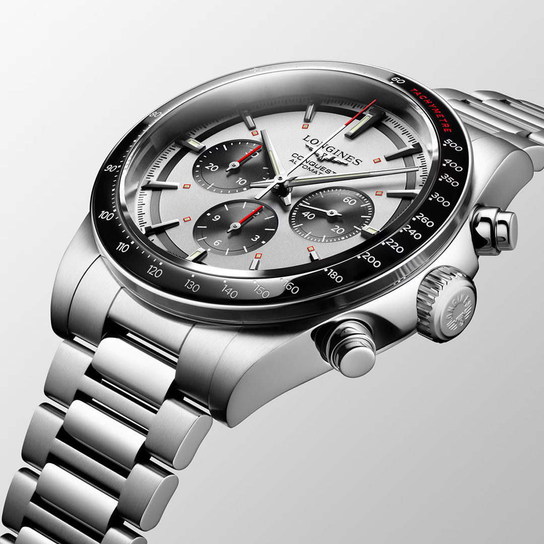 A close-up of the Longines Conquest Chronograph Automatic 42mm Watch, featuring a stainless steel case, silver dial with three subdials, black bezel, and red accents, showcased at an angle on a light gray background.
