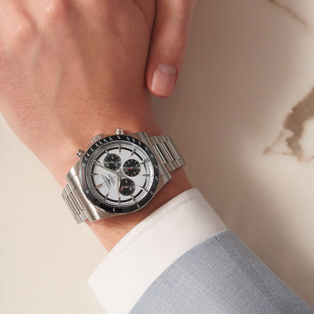 Wearing a Longines Conquest Chronograph Automatic 42mm watch with a white dial and three subdials, someone in a light blue suit jacket and white shirt rests their hand on a marble surface.