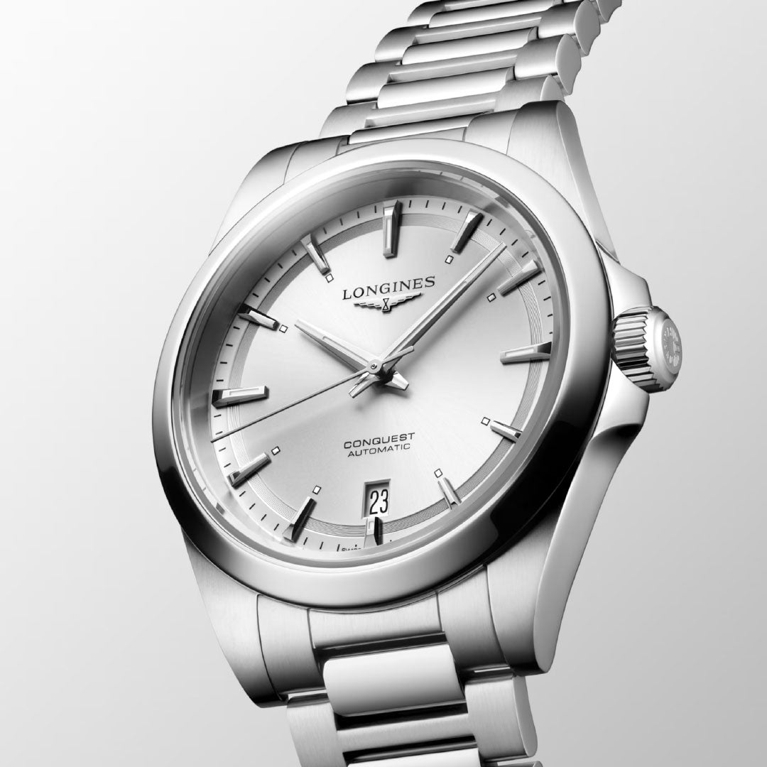 A close-up of the Longines Conquest Automatic 38mm Watch highlights its sporty elegance with a silver dial, silver hour markers, date at 6 o'clock, and stainless steel bracelet against a light background.