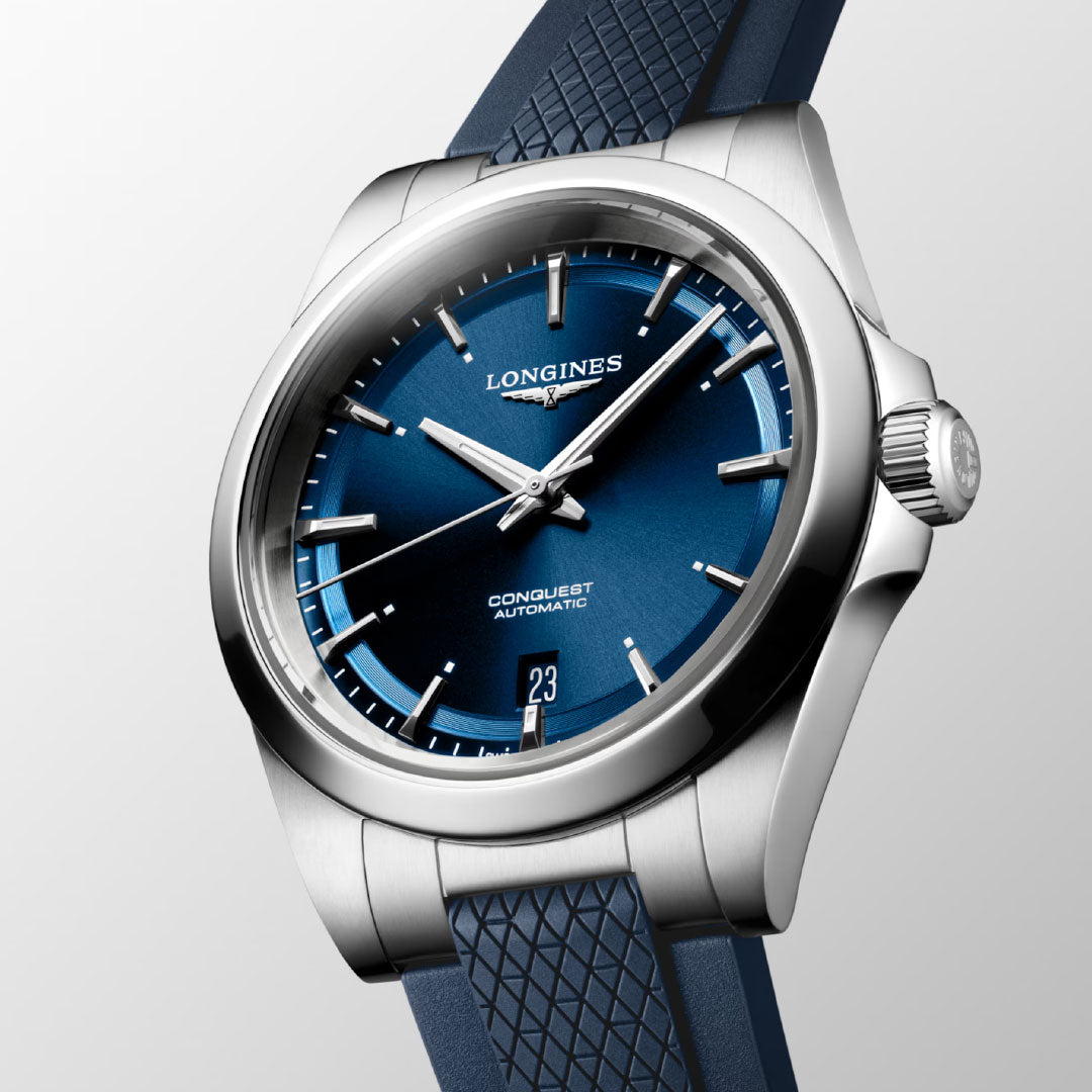 A close-up of the Longines Conquest Automatic 38mm Watch showcases its sporty elegance with a blue textured strap, blue dial, silver hour markers, and a date window at 6 o’clock, set against a light gradient background.