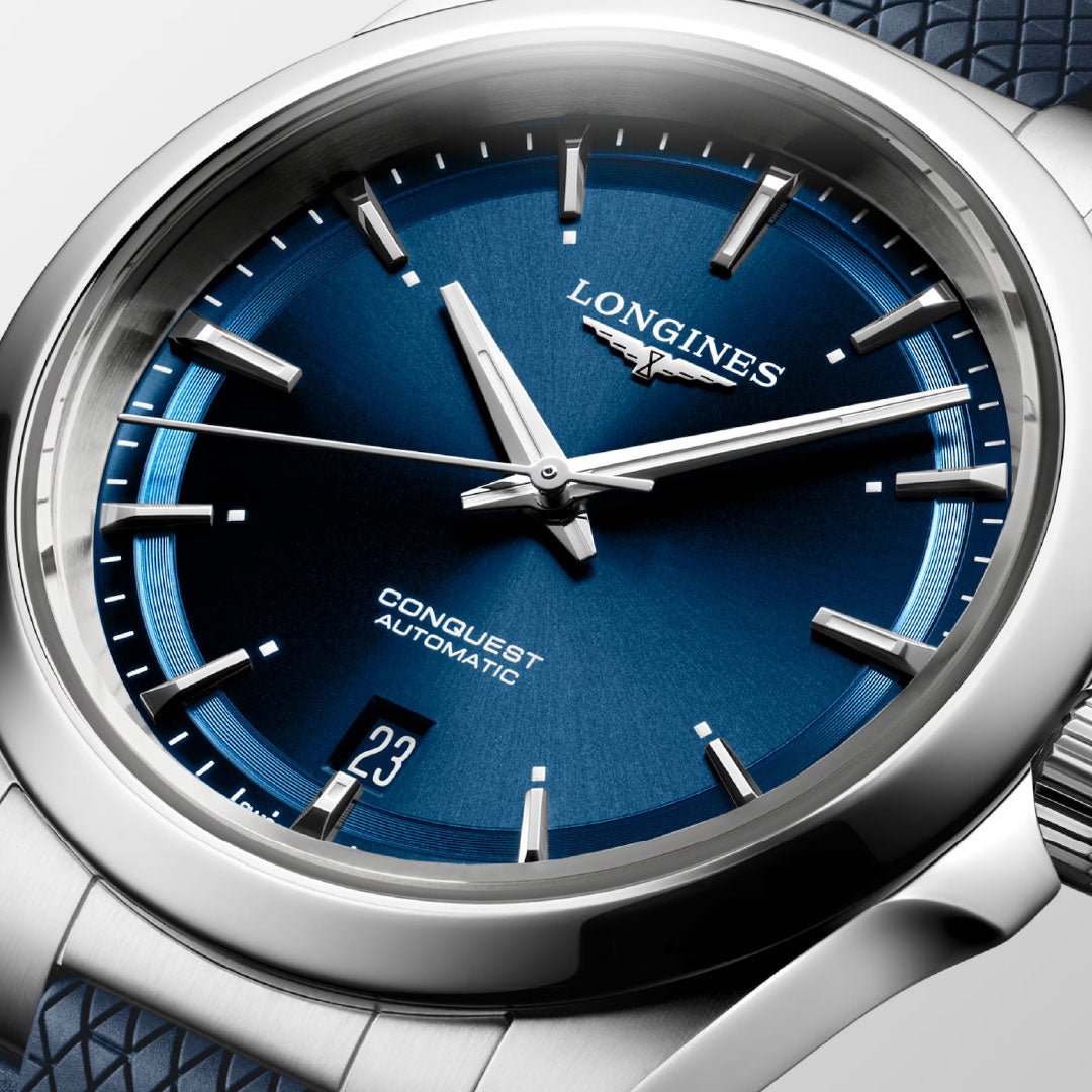 Close-up of the Longines Conquest Automatic 38mm, featuring a blue textured dial, silver markers and hands, date window at 6 o’clock, and partial view of its stainless steel case with a blue strap.