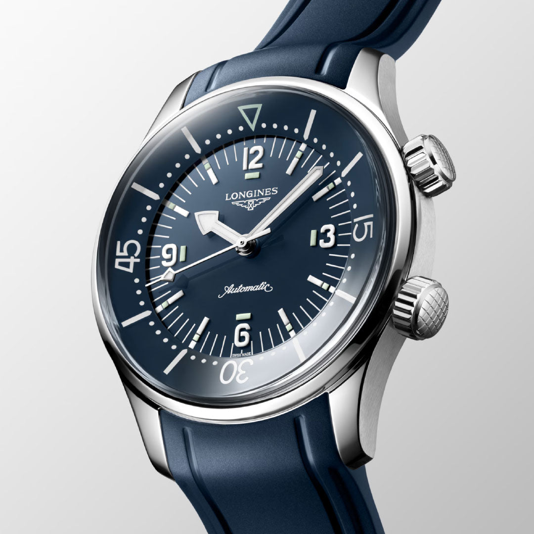 A close-up of the Longines Legend Diver Automatic 39mm Watch shows a blue dial with white markers, blue strap, silver-tone case, two crowns, and an internal rotating bezel by Longines, set against a light gray background.