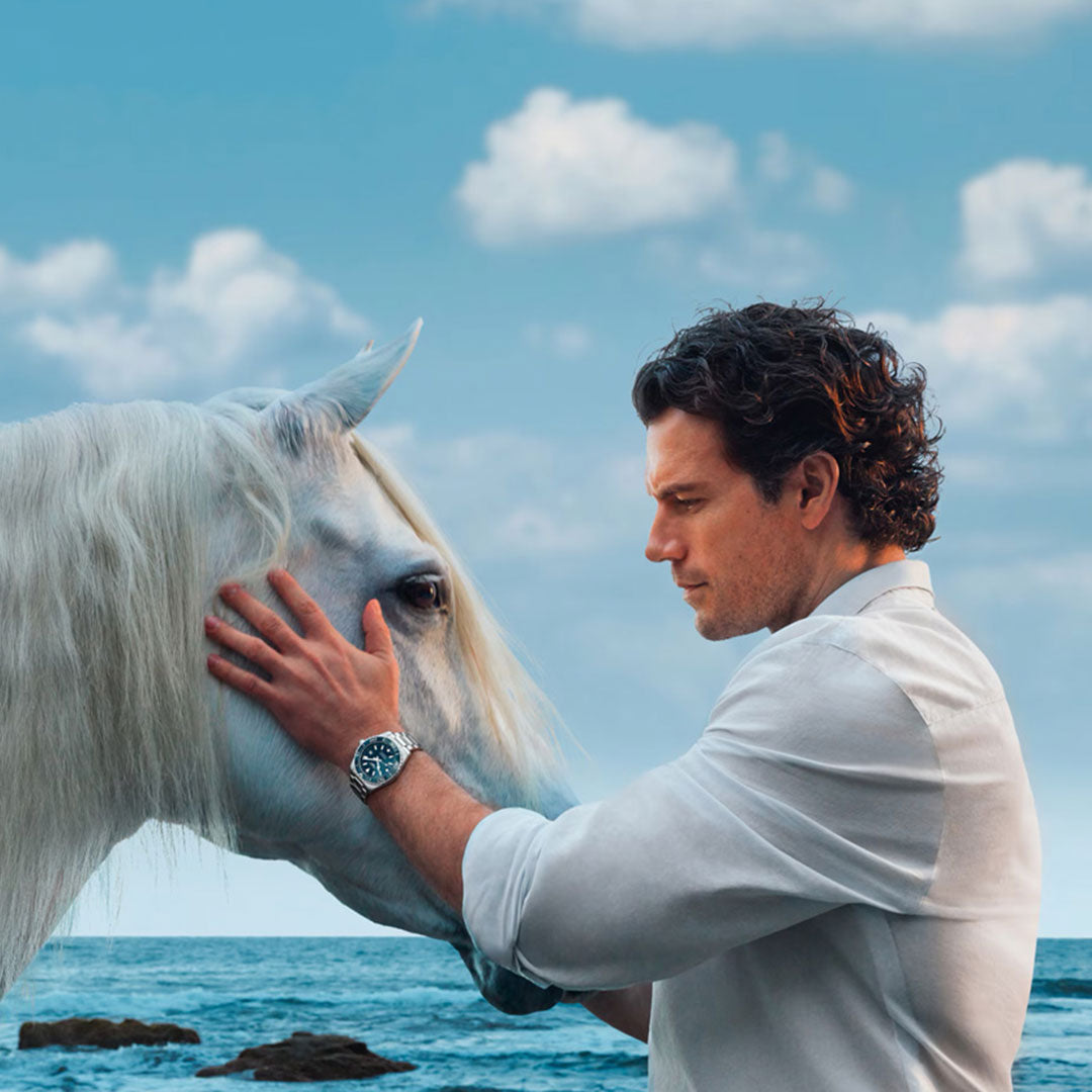 A man in a white shirt gently touches a white horse’s face by the ocean under a blue sky—an image of timeless elegance, echoed by the water-resistant Longines Hydroconquest Automatic 42mm Watch.