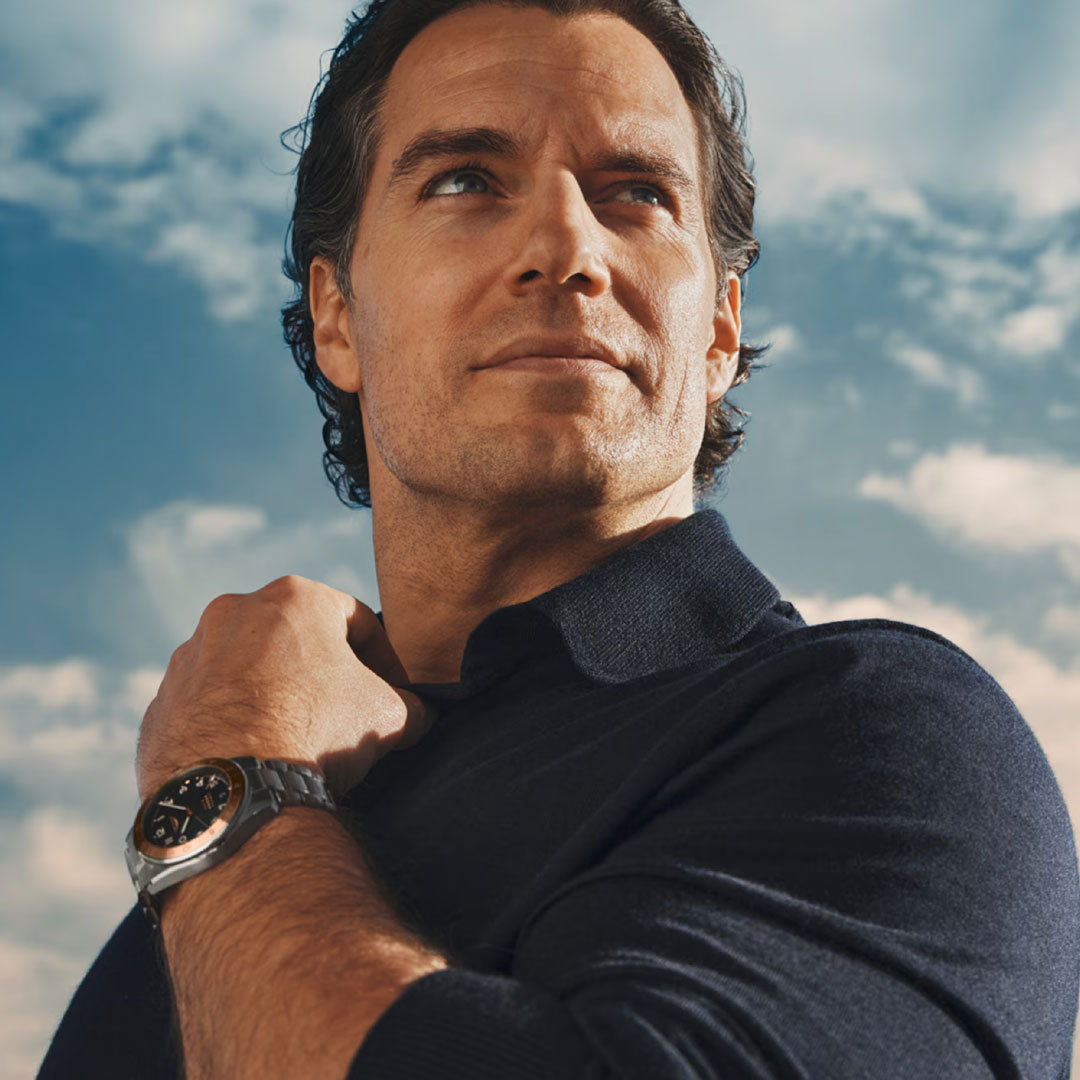 A man with wavy brown hair in a navy shirt looks into the distance, holding his collar. He wears a Longines Spirit Zulu Time 1925 Automatic 39mm Watch by Longines, standing against a blue sky dotted with clouds.