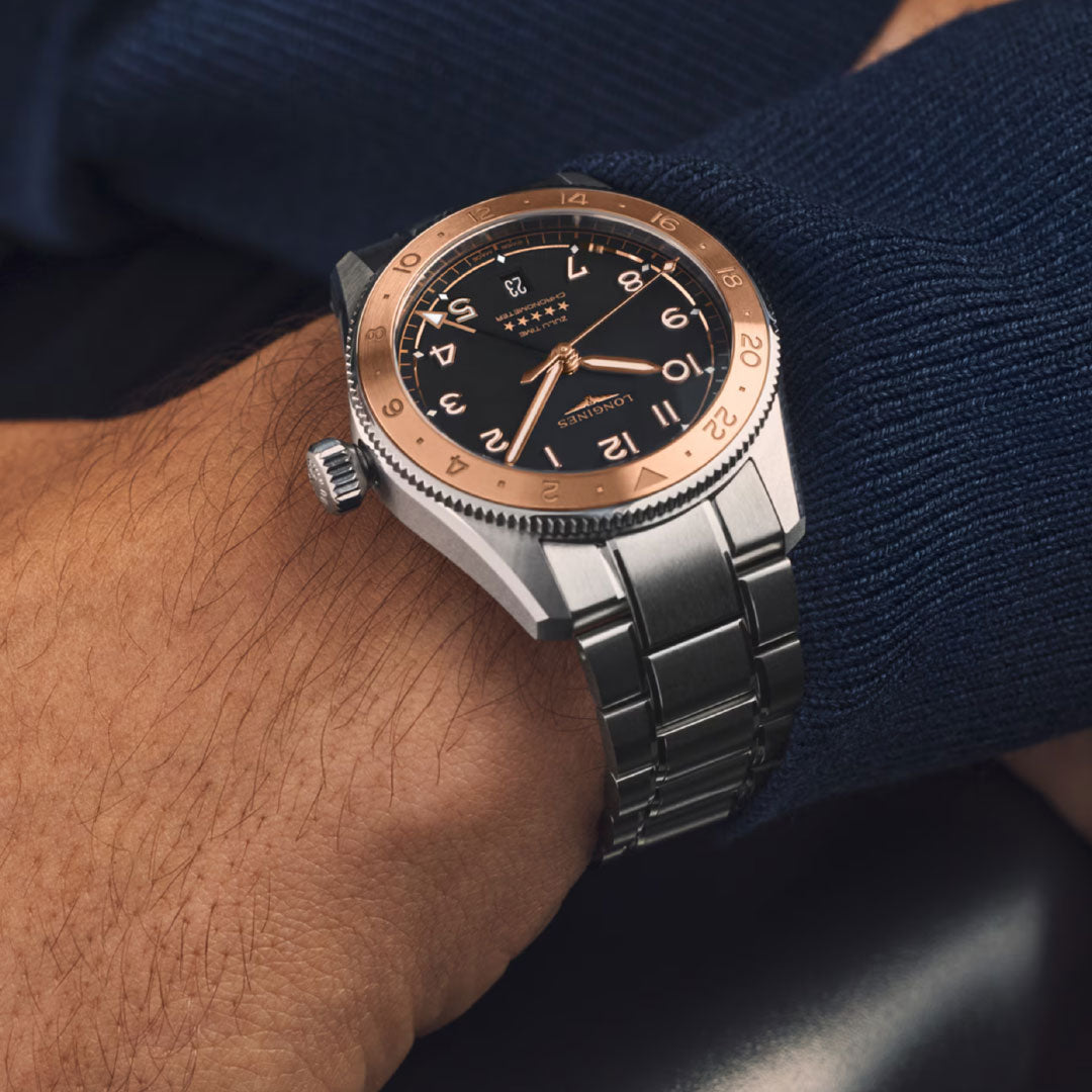 A close-up of a wrist wearing the Longines Spirit Zulu Time 1925 Automatic 39mm watch in silver and rose gold with a black dial and Arabic numerals, styled with a navy blue sweater.