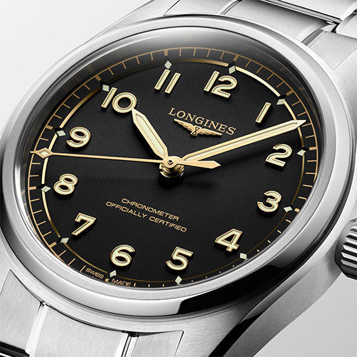 A close-up of the Longines Spirit Pilot Automatic 39mm watch shows a silver case, black dial with gold-tone Arabic numerals and hands, "Chronometer Officially Certified" above 6 o’clock, and a metal bracelet.