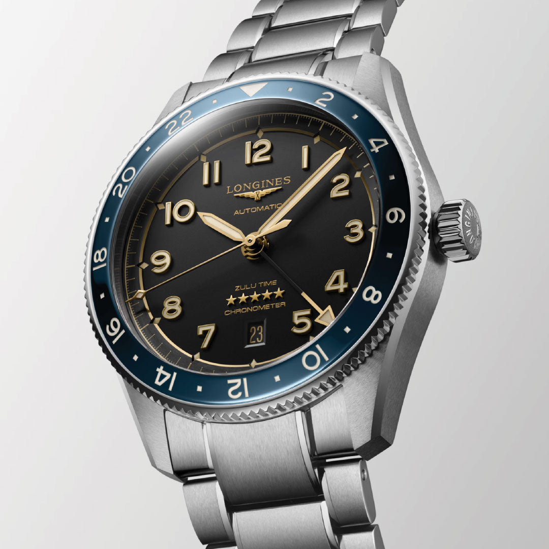 A close-up of the Longines Spirit Zulu Time Automatic 42mm Watch, featuring a stainless steel bracelet, black dial with gold-tone numerals, blue bezel, date at 6 o’clock, chronometer certification, dual time zones, and signature five stars.