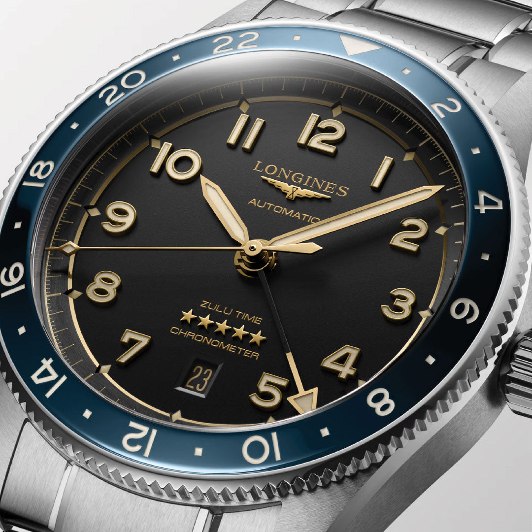 Close-up of the Longines Spirit Zulu Time Automatic 42mm Watch featuring a black dial, gold numerals and hands, silver case, blue bezel, date at 6 o’clock, and "ZULU TIME" text above stars.