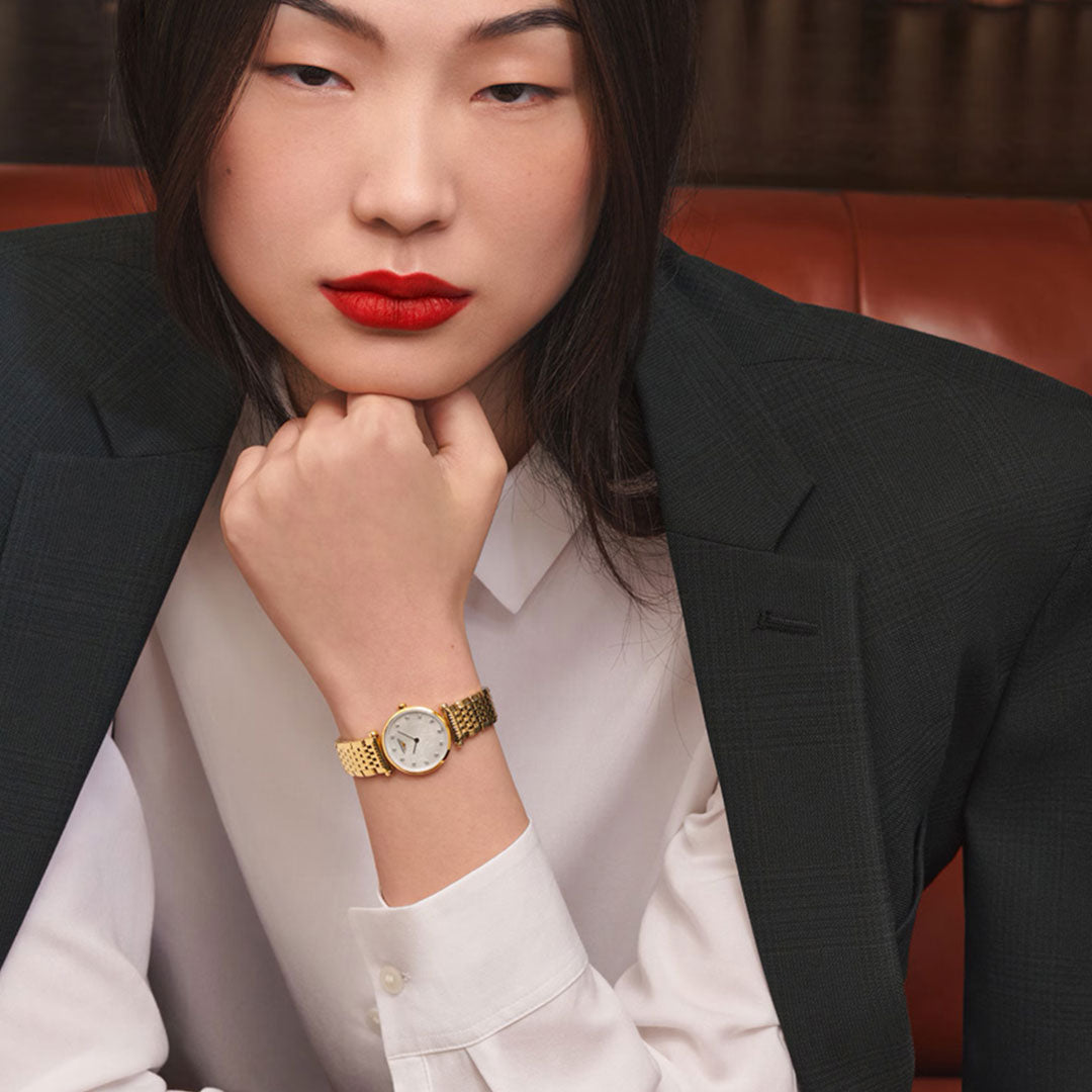 A person with long dark hair, red lipstick, and a serious expression sits on a brown leather seat, exuding classic elegance in a dark blazer and white shirt while wearing the Longines La Grande Classique Quartz 24mm watch.
