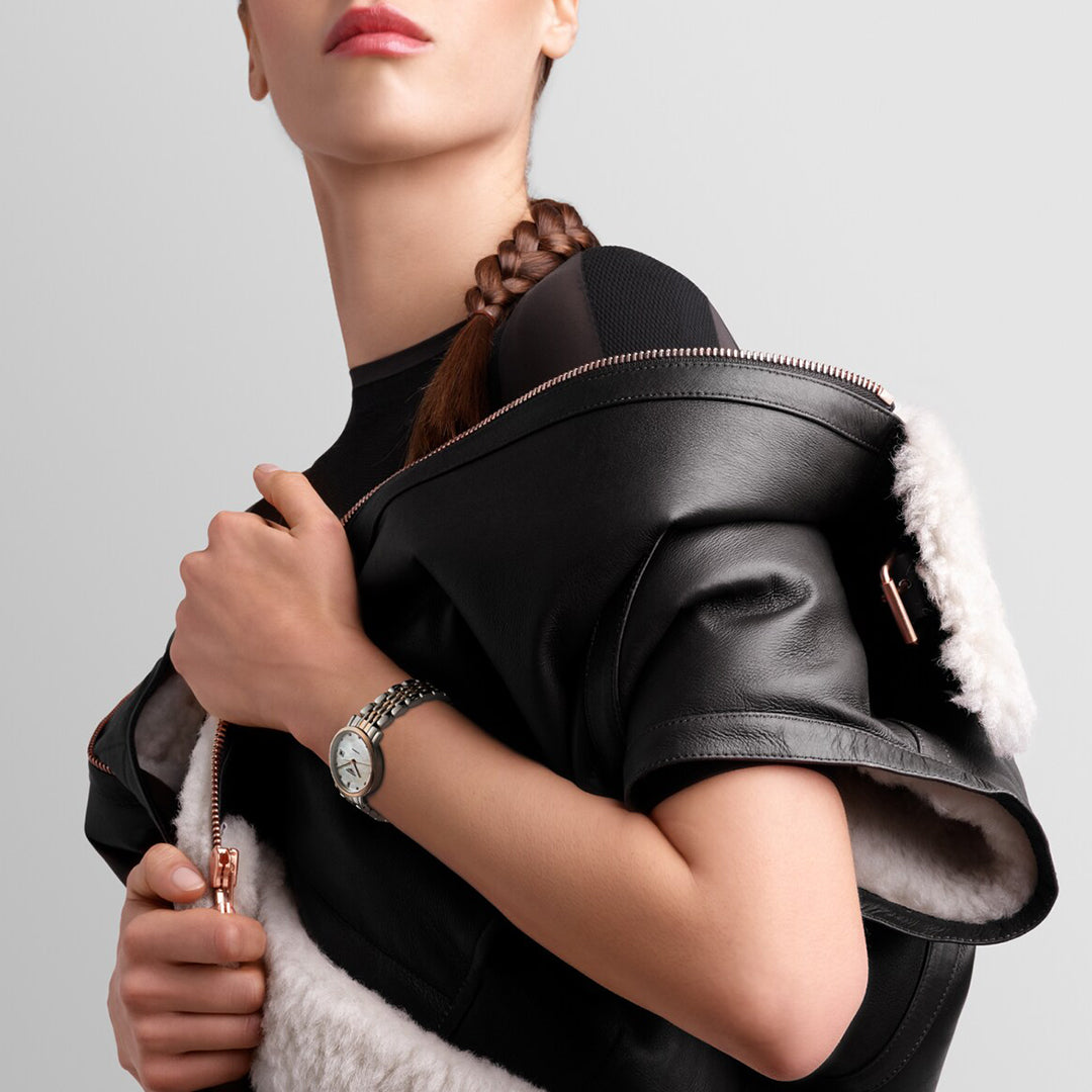 A person with a braid carries a worn black leather jacket with white fur lining on their shoulder, wearing a black top and showcasing the Longines Elegant Collection Automatic 29mm Watch, epitomizing timeless elegance against a plain gray background.