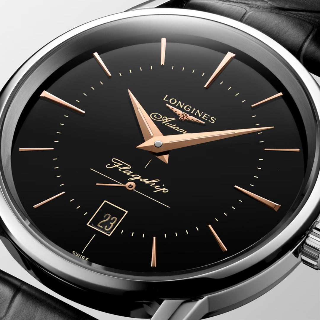A close-up of the Longines Flagship Heritage Automatic 38.5mm Watch, featuring a black dial with gold markers and hands, a date window at 6 o’clock, the Longines logo, “Automatic” text, and a black leather strap.