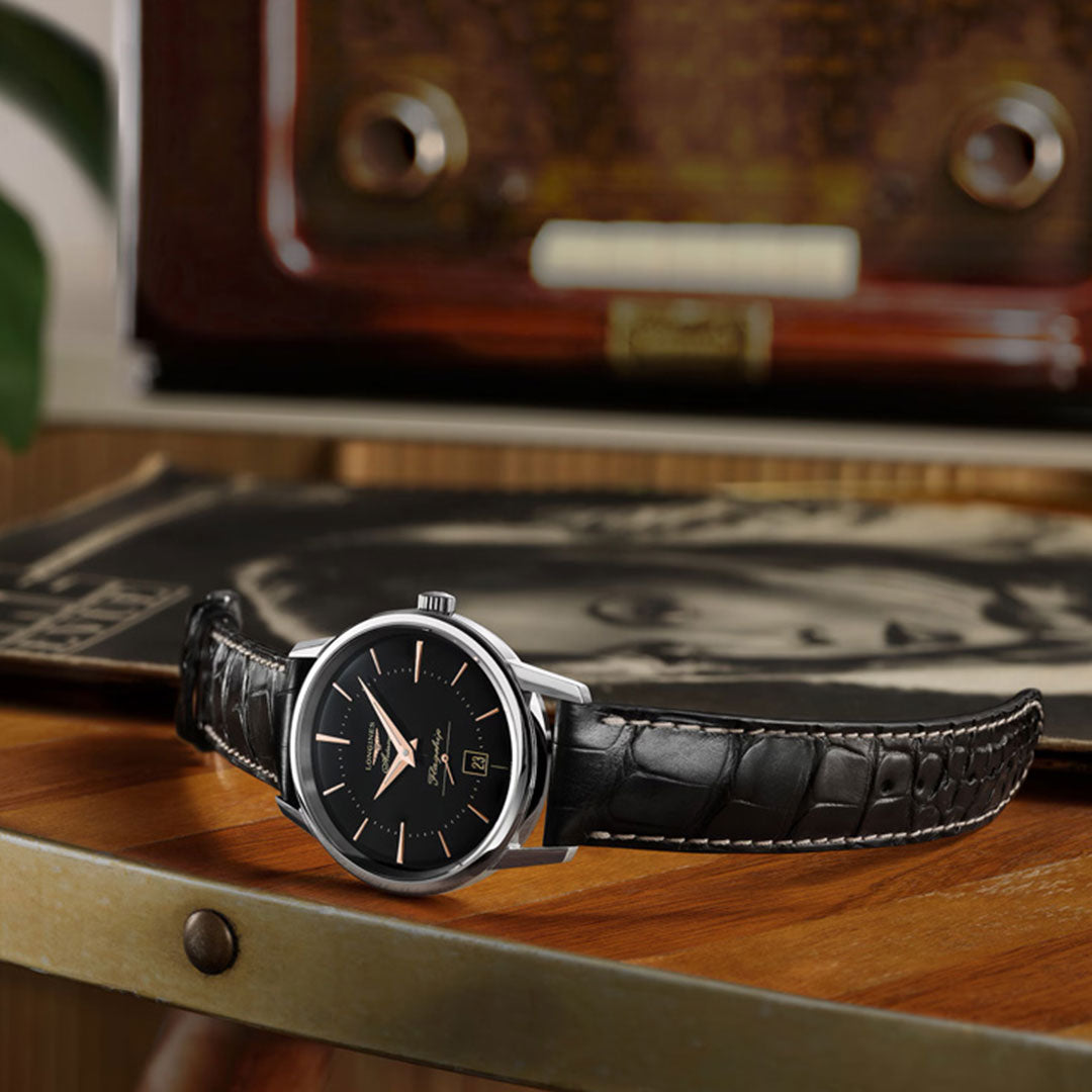 The Longines Flagship Heritage Automatic 38.5mm Watch by Longines, featuring a black leather strap and silver case, rests on a wooden table with a vintage radio and vinyl record softly blurred in the background, evoking timeless elegance.