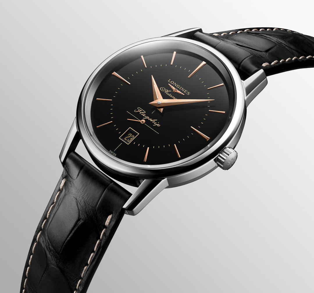 A close-up of the Longines Flagship Heritage Automatic 38.5mm Watch, featuring a black leather strap, black dial, gold hands and markers, and a date window at 6 o’clock—an elegant classic by Longines on a light gradient background.