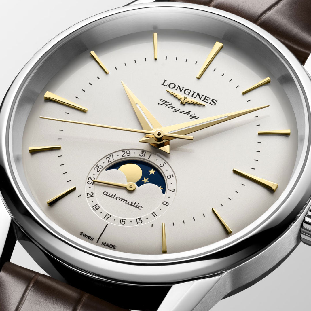 Close-up of the Longines Flagship Heritage Moonphase Automatic 38.5mm Watch featuring a silver dial, gold hour markers, moon phase display, brown leather strap, and an enamel caravel medallion on the case back.